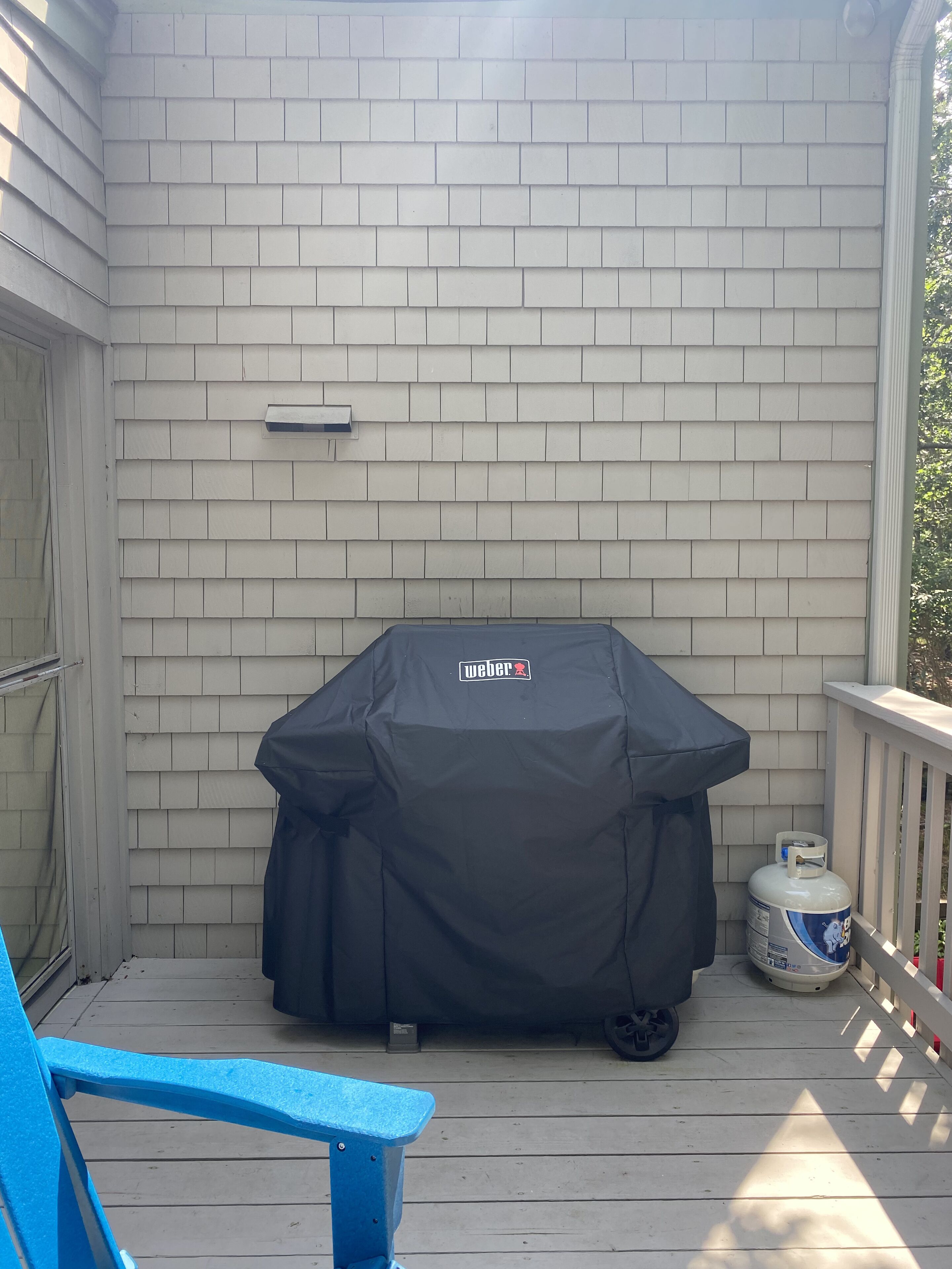 A covered grill on a wooden deck with a white exterior wall and a blue chair.