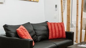 Un sofá de cuero negro con dos almohadas rojas, una mesita negra pequeña y un separador de bambú al fondo.