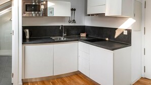 Una cocina compacta con armarios blancos, encimera negra, microondas, hervidor y lavabo.
