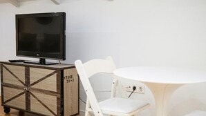 Una habitación con un televisor sobre un mueble de madera, sillas blancas y una mesita pequeña blanca.