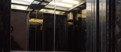 An elevator interior with a reflective wall, a patterned carpet, and a ceiling with recessed lighting.