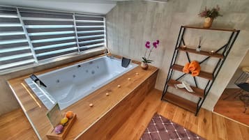 A modern bathroom with a wooden vanity, a large bathtub, a wooden shelf with towels, and a window with blinds.