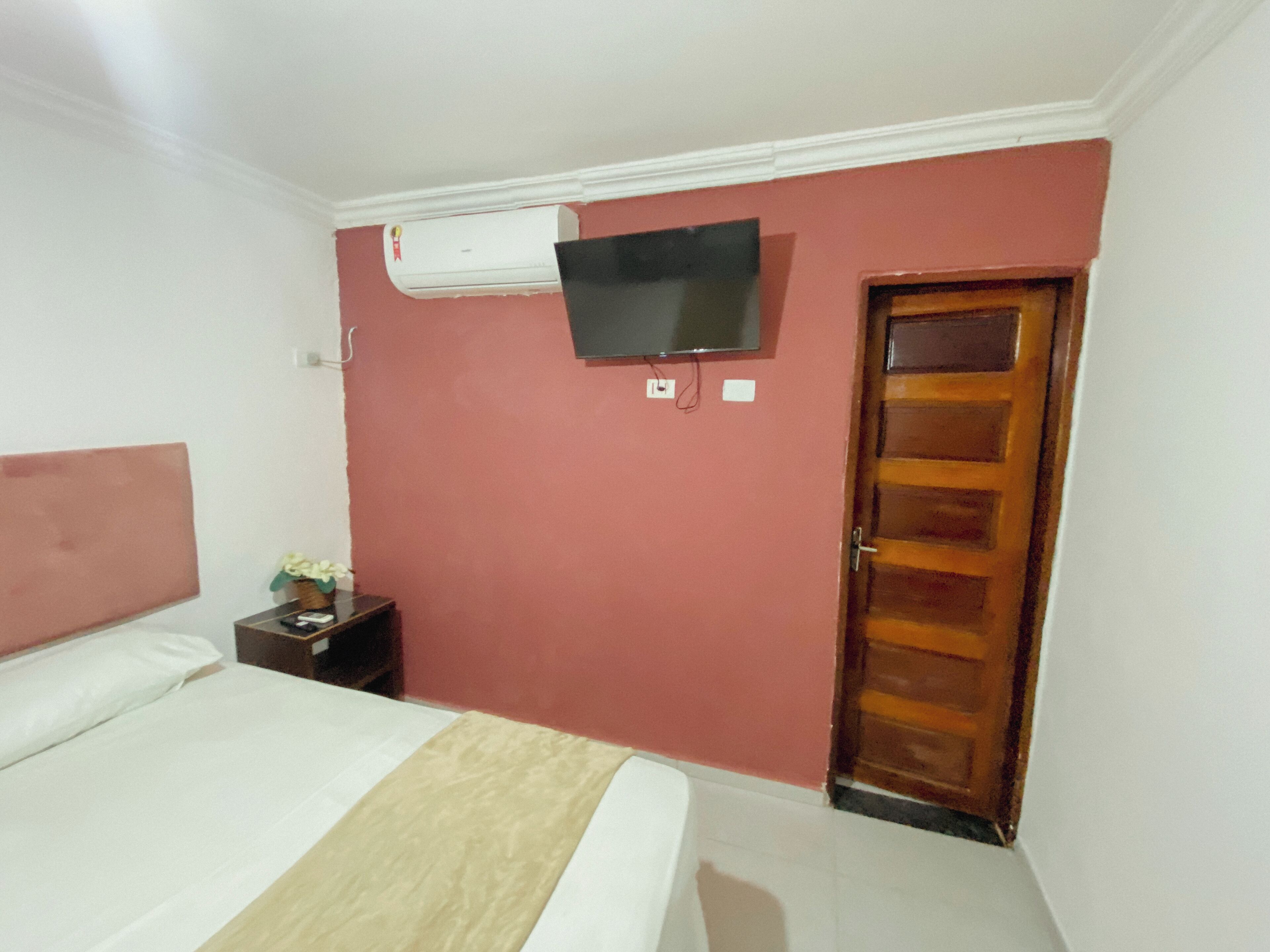 Una habitación de hotel con una cama, un televisor montado en la pared, un equipo de aire acondicionado y una puerta de madera.