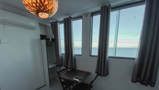 A compact room with a dining table and chairs, a hanging light fixture, and large windows with curtains. - Hotel 5 Sóis (Olinda)