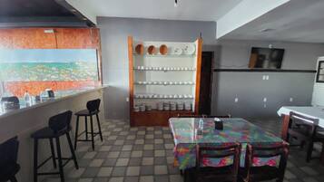A dining area with a table and chairs, a bar counter with stools, and a shelf with plates and cups.