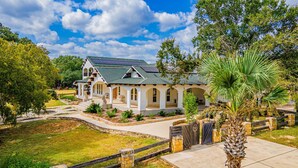A white building with a green roof and solar panels, surrounded by a well-maintained garden and a stone fence. - Casa Blanca Main House, Riverview, 10 Beds, Group-Friendly (Kerr County)