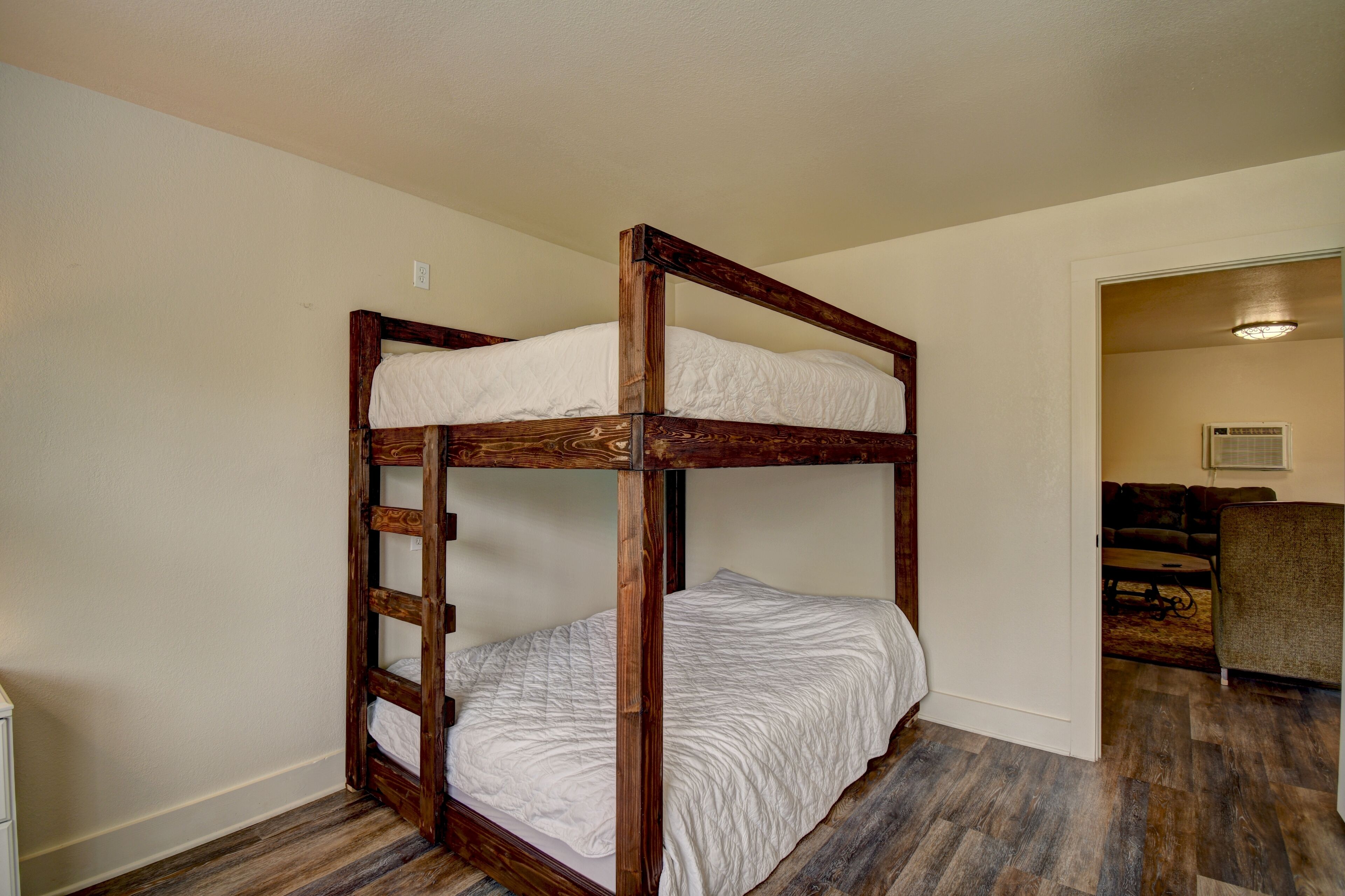 A bunk bed with wooden railings and white bedding in a room with wooden flooring and a door leading to another room.