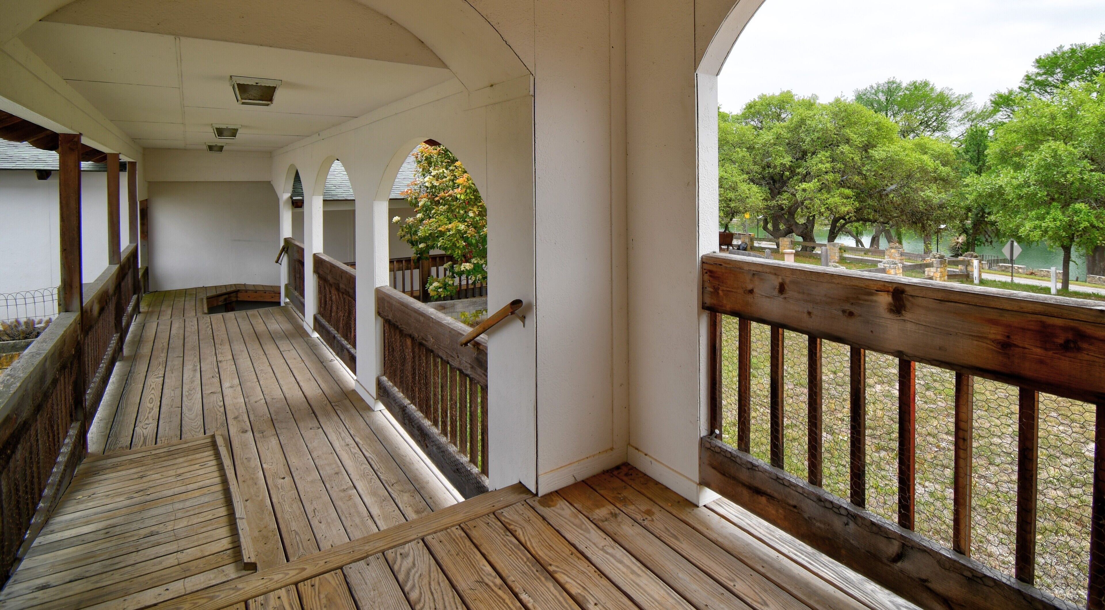 A covered wooden deck with a railing, arched columns, and a view of a park with trees and a fence.