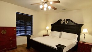 Une chambre avec un plancher en bois, un lit recouvert de draps blancs, deux tables de chevet avec des lampes, un ventilateur de plafond et une fenêtre avec des stores.