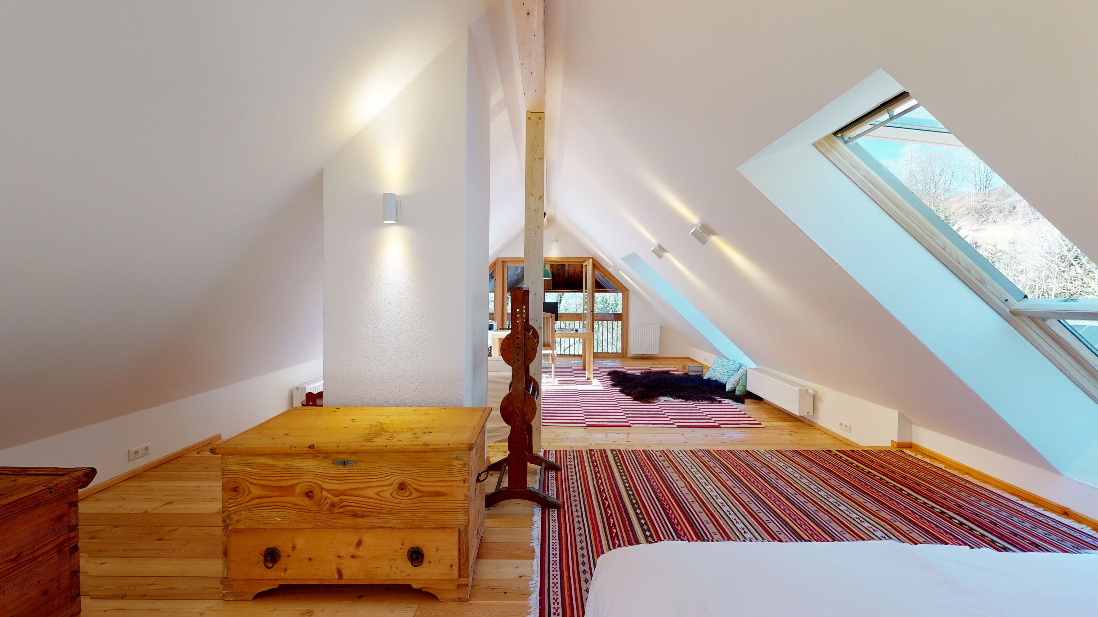 A high-ceilinged attic bedroom with a sloped roof, wooden flooring, a large window, and a view of trees outside.