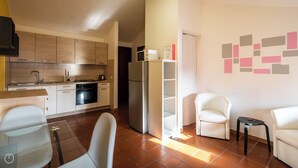 Una cucina compatta con elettrodomestici moderni, una zona pranzo con tavolo e sedie bianche, e un soggiorno con divano e un piccolo tavolino.