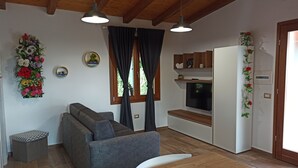 Un espacio habitable moderno con un sofá gris, vigas de techo de madera, una mesa de comedor con un tarro de vidrio y una zona de cocina con televisión y armarios.