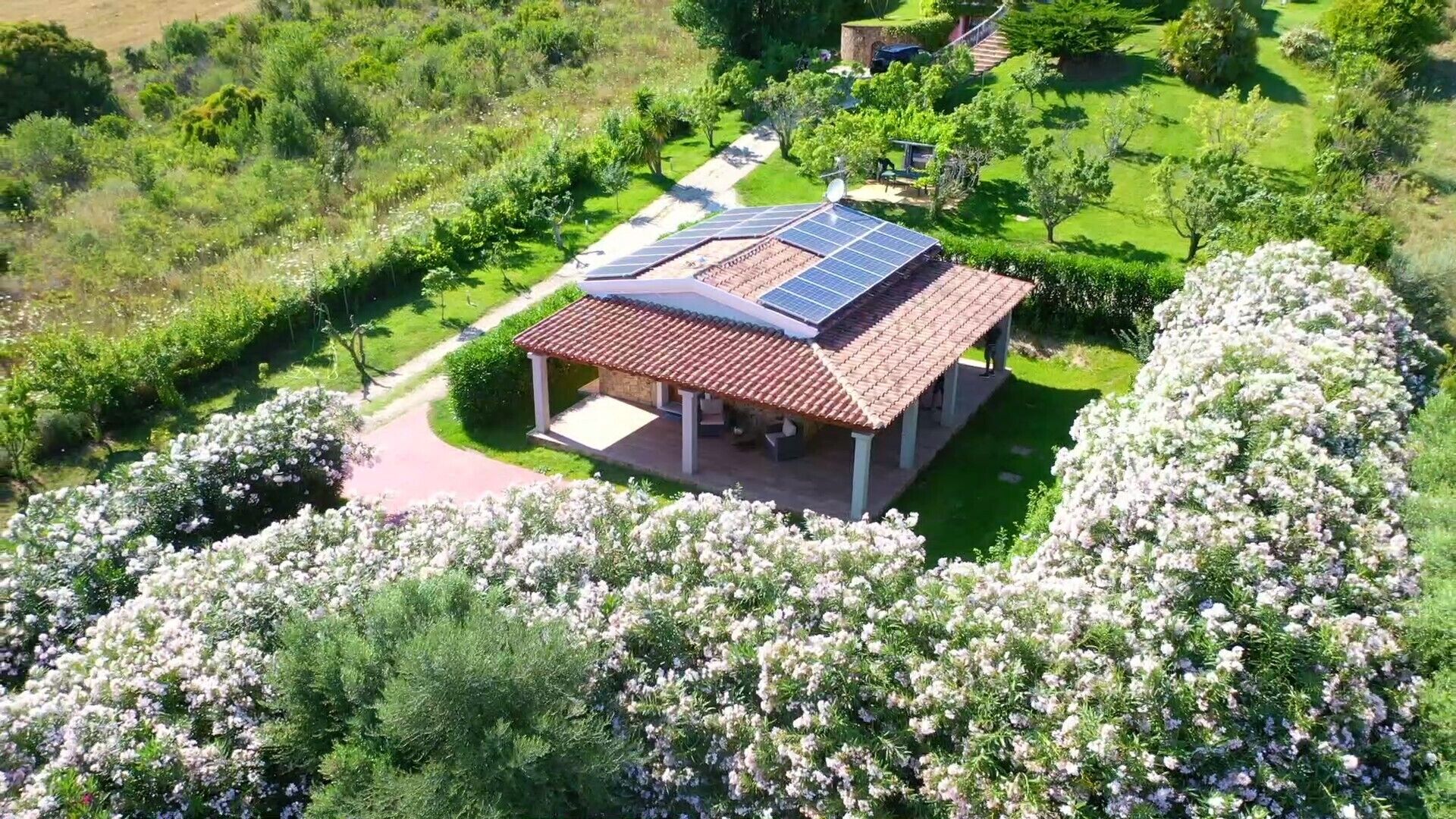 Un pabellón con techo de tejas y paneles solares, rodeado de exuberante vegetación y arbustos florecientes.