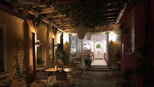 An outdoor café area with wooden tables and chairs, surrounded by plants and a brick pathway. - Weingut Manuel Sauer (Nordheim a. Main)
