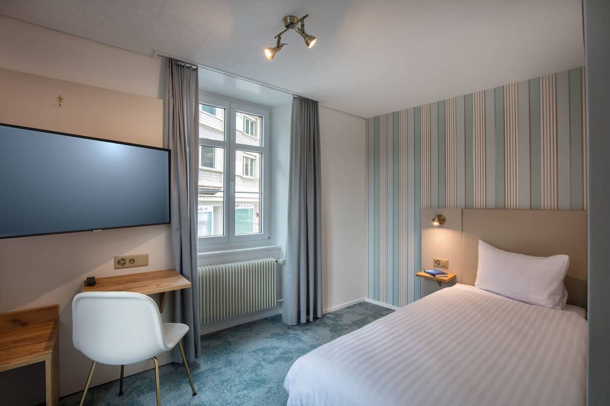 A hotel room with a bed, a desk, a chair, a TV, a window with curtains, and a striped wall.