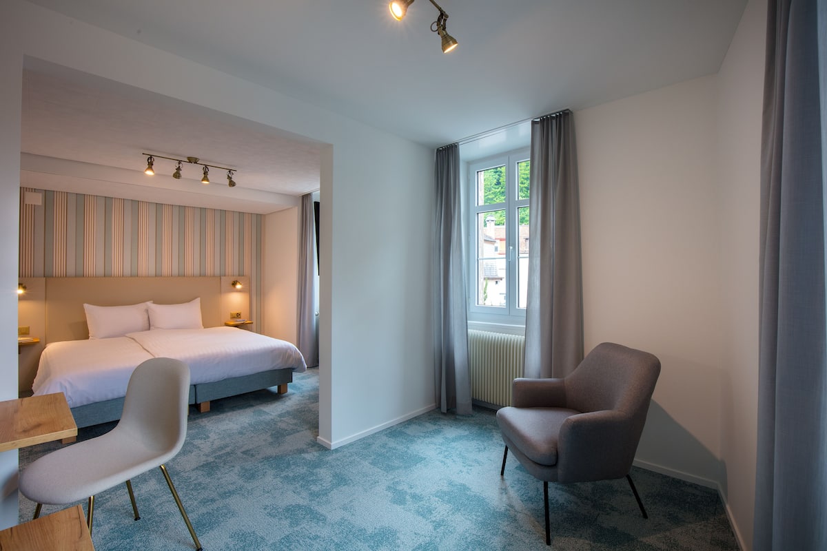 A hotel room with a bed, a chair, a table, and a window with curtains.