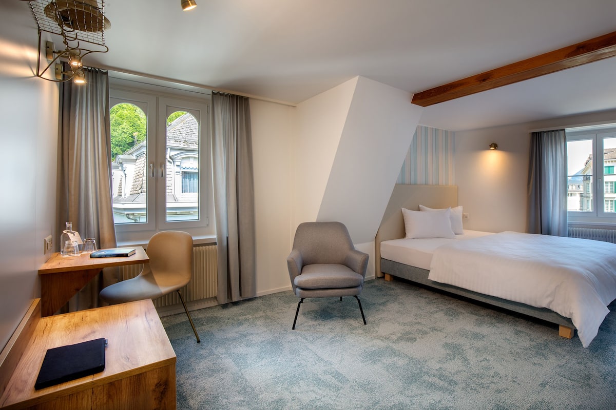 A hotel room with a bed, a desk with a chair, a window with curtains, and a view of buildings outside.