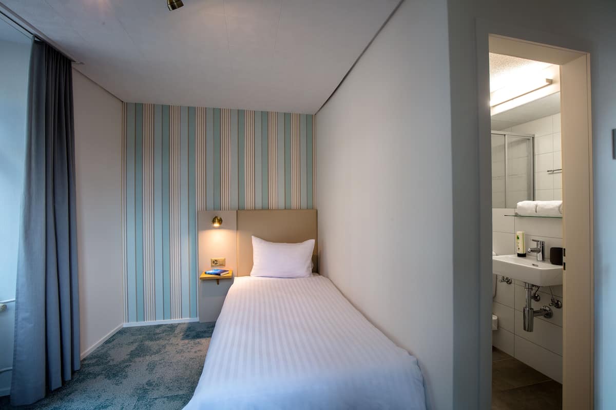 A hotel room with a single bed, a nightstand with a lamp, a striped wall, and a bathroom visible through an open door.
