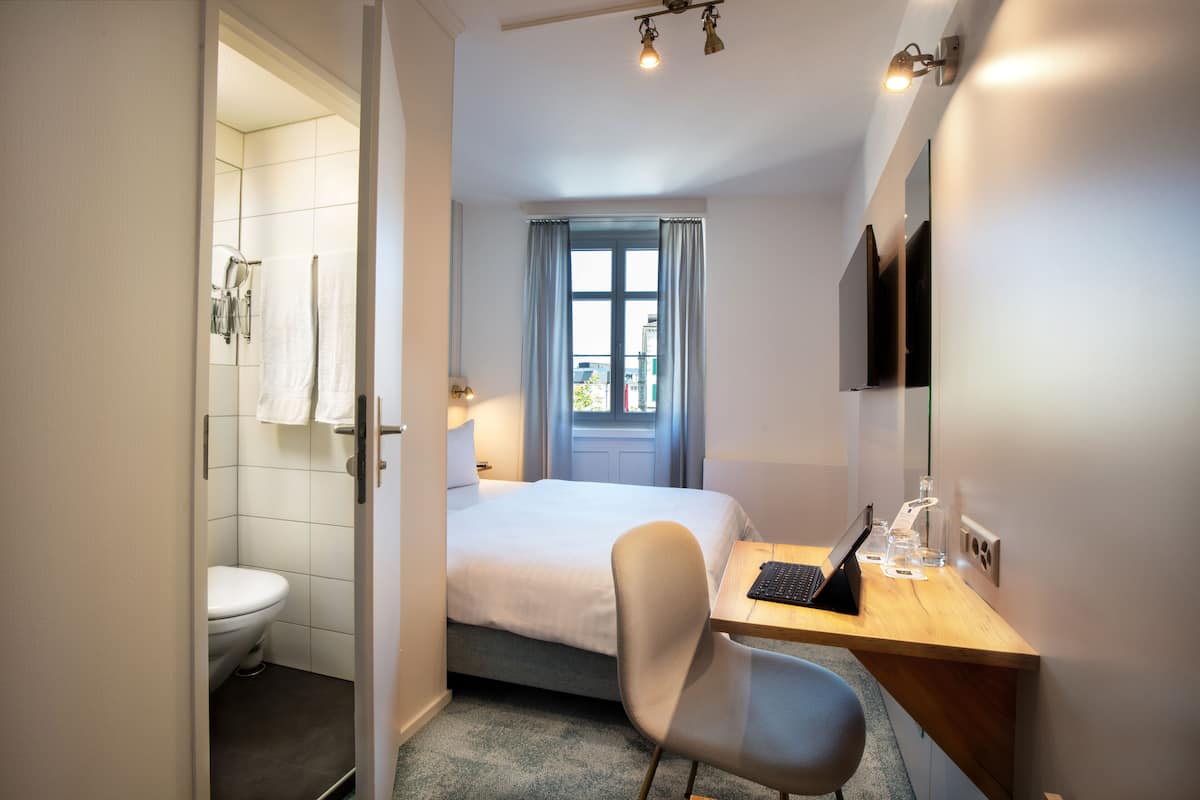 A hotel room with a bed, a desk with a laptop, a chair, a TV, and a window with curtains.