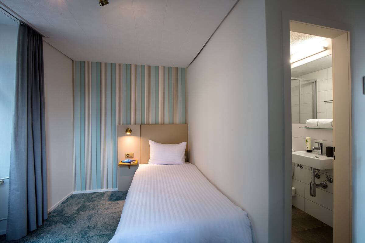 A hotel room with a bed, a nightstand with a lamp, a bathroom with a sink and mirror, and striped wallpaper.