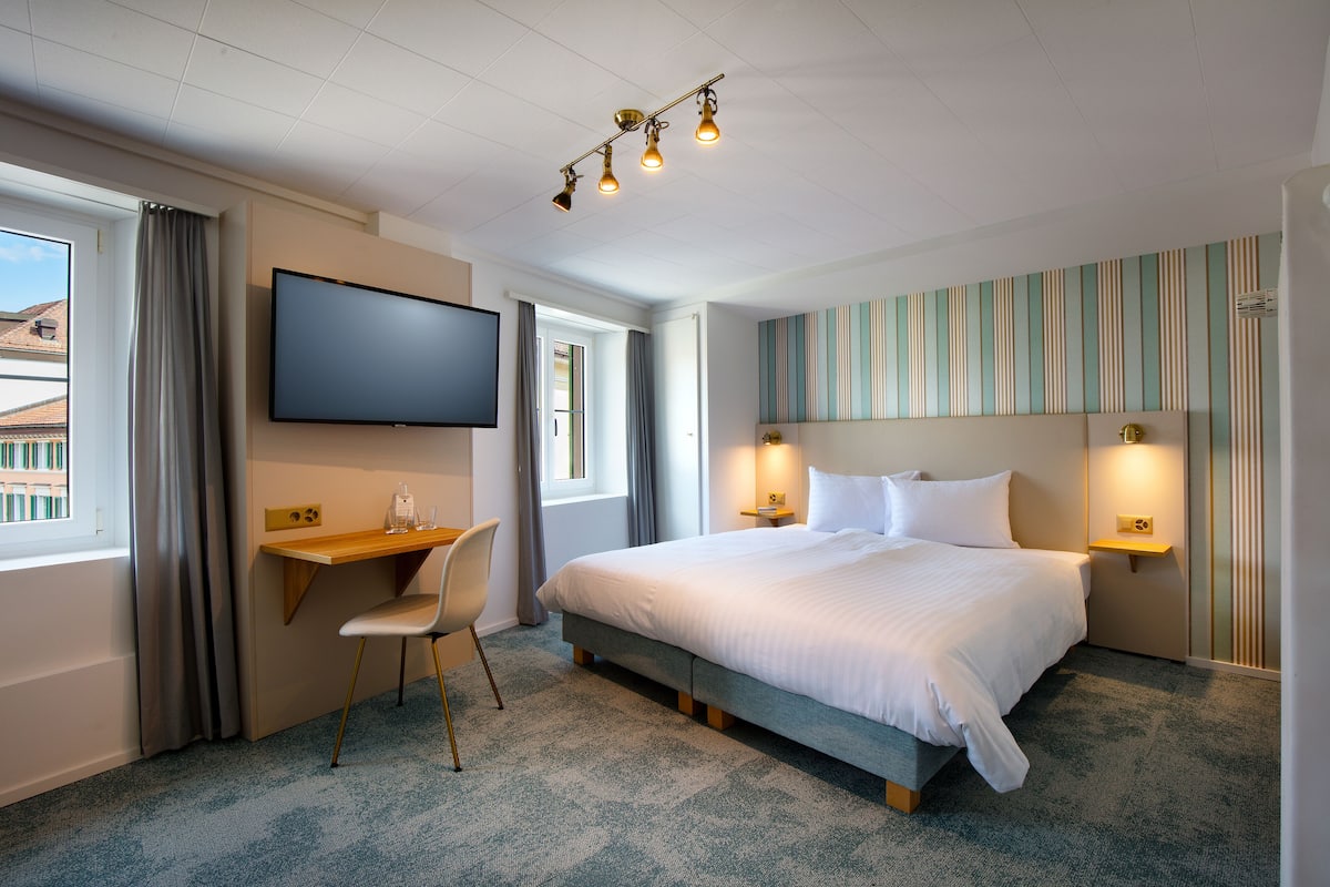 A hotel room with a bed, a desk, a chair, a TV, and a window with curtains.