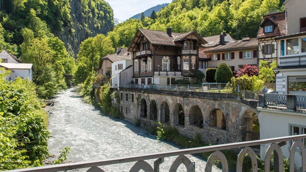 Un pueblo a orillas del río con arquitectura tradicional, un puente de piedra y abundante vegetación.