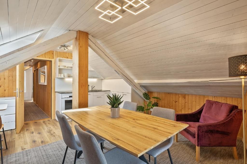Ein modernes Loft mit Holzbalkendecke, einem Essbereich mit Holztisch und -stühlen, einem roten Samtstuhl und einer Küche im Hintergrund.
