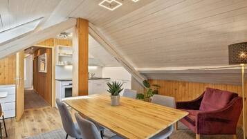Ein modernes Loft mit Holzbalkendecke, einem Essbereich mit Holztisch und -stühlen, einem roten Samtstuhl und einer Küche im Hintergrund.
