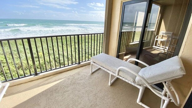 Un balcon avec vue sur l’océan, un fauteuil de relaxation blanc et une porte vitrée donnant sur un espace ombragé avec une table et des chaises.