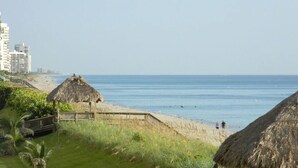 Une plage avec des cabanes aux toits de chaume, une promenade en bois et une vue lointaine sur des bâtiments.