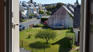 Vue d’une rue résidentielle à travers une fenêtre, avec des maisons, un petit espace vert et un véhicule garé.