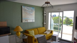 Un salon avec un canapé jaune, une petite table sur laquelle se trouve une lampe, un fauteuil et un tableau représentant une scène de plage.