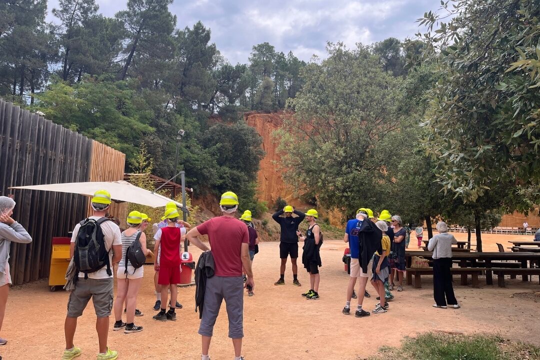 A group of people standing outdoors, some wearing yellow hats, near a wooden structure and a rocky cliff.