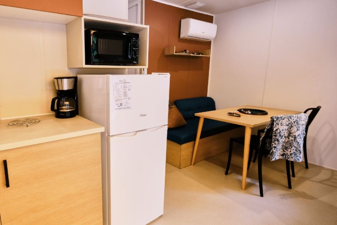 Eine kompakte Küchenzeile mit Mikrowelle, Kaffeemaschine und Kühlschrank. Ein kleiner Essbereich mit Tisch und einem Stuhl ist vorhanden.
