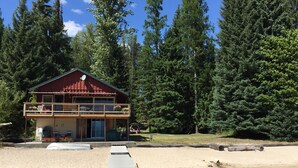 A two-story cabin with a balcony, a sandy beach in the foreground, and a forest in the background. - Priest Lake BEACHFRONT w 2 slip Dock (Priest Lake Idaho)