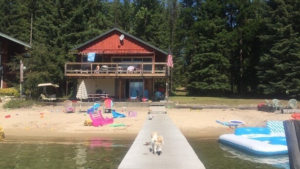 A wooden dock leading to a house with a red roof, surrounded by a sandy beach and inflatable pool. - Priest Lake BEACHFRONT w 2 slip Dock (Priest Lake Idaho)