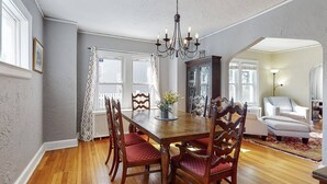 A dining area with a wooden table, red cushioned chairs, a chandelier, and a view of a living room with a white armchair and a lamp. - Belltown Retreat - Shopping, Dining, Pet Friendly (Stamford)