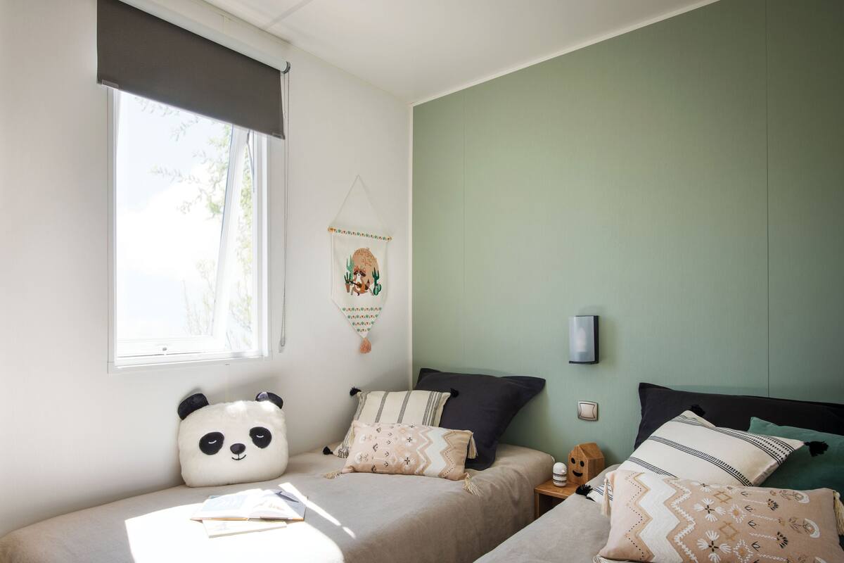 Une chambre avec deux lits, une fenêtre avec un store, une lampe fixée au mur et un élément décoratif mural.