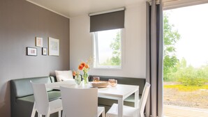 A modern living room with a green sofa, white chairs, a dining table, and a window with a view of greenery. - Camping Pascalounet (Martigues)