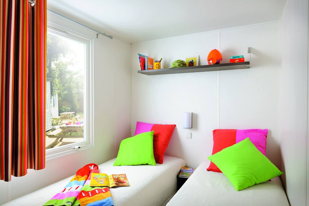 Une petite pièce lumineuse, avec deux lits, des coussins colorés, une étagère remplie de livres et un casque, et une fenêtre avec des rideaux.