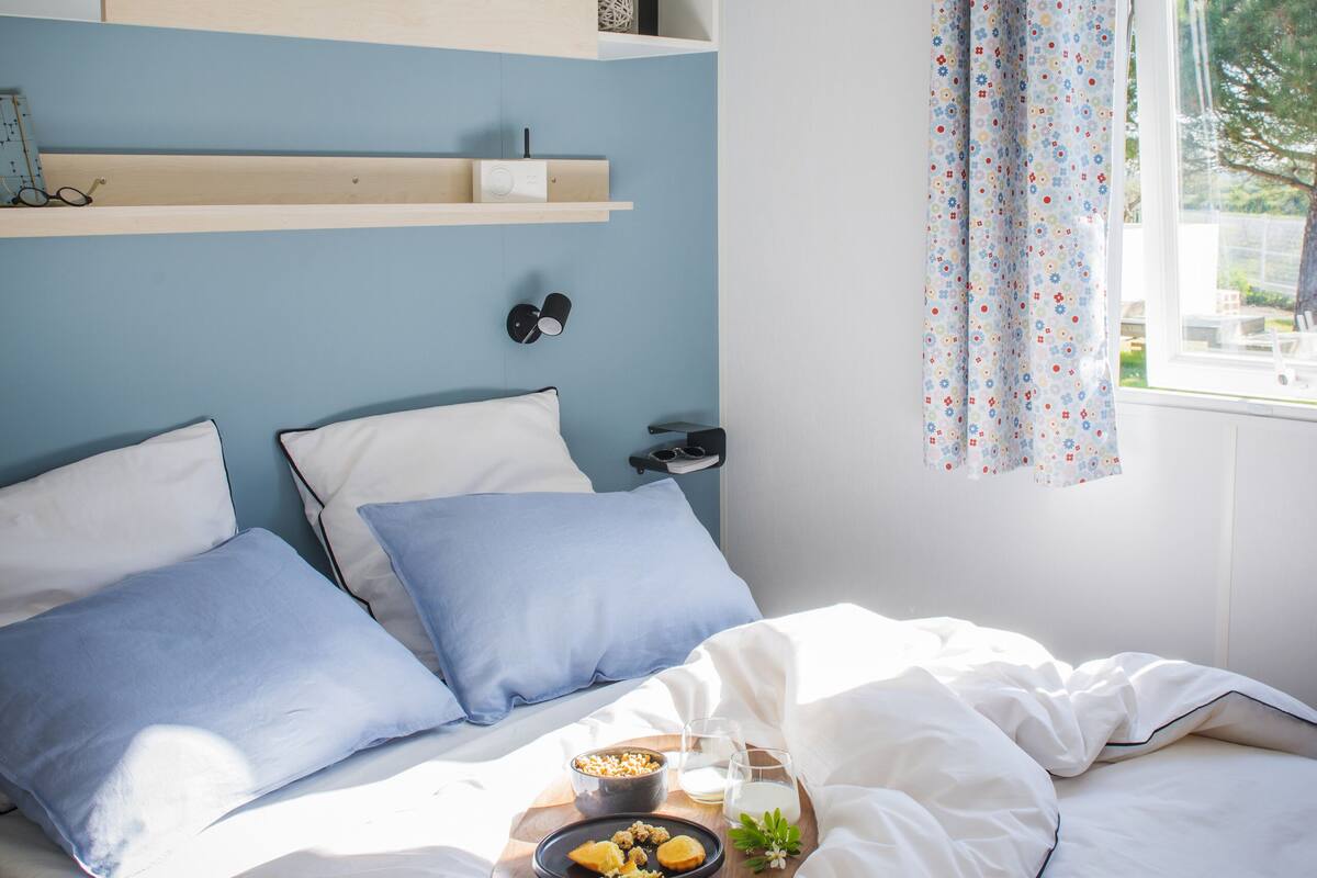Une chambre à coucher avec un lit, des oreillers, une table de chevet avec une lampe, une fenêtre avec des rideaux et une étagère au-dessus du lit.