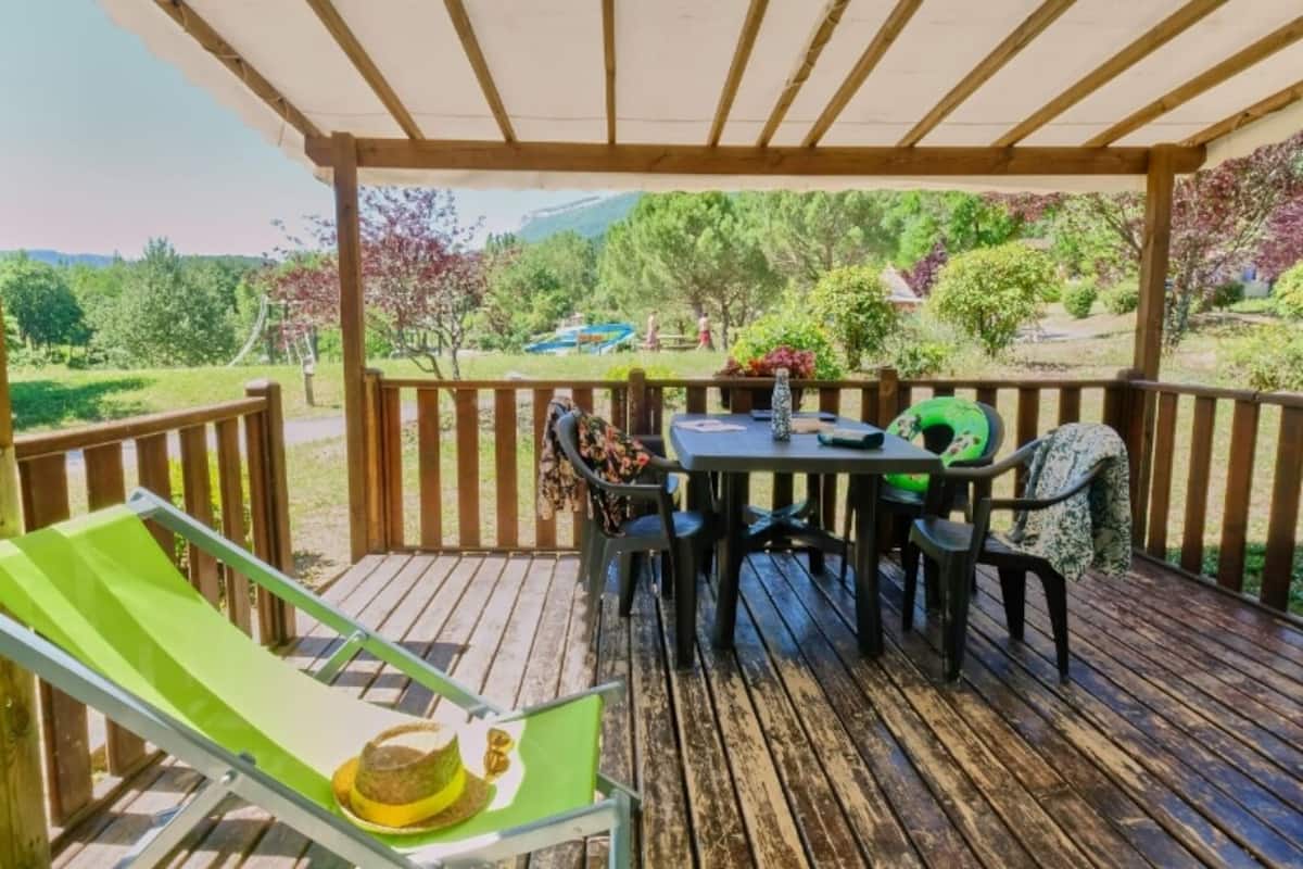 Eine Holzterrasse mit Tisch und Stühlen, ein grüner Liegestuhl und Blick auf einen Park und Berge.