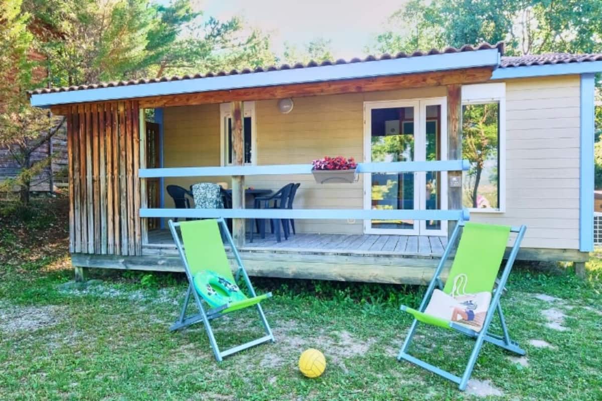 Ein kleines, eingeschossiges Haus mit Veranda, zwei grüne Rasensessel und ein gelber Ball auf dem Rasen.
