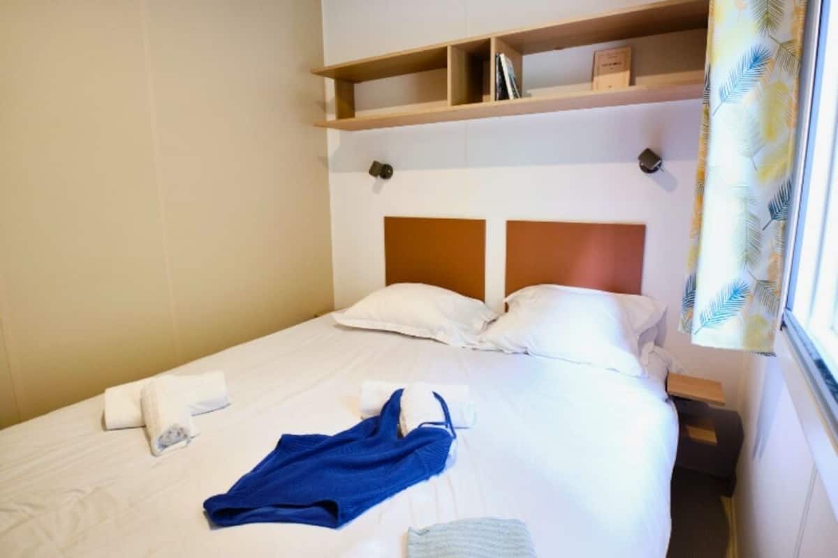 A hotel room with a bed, white linens, a blue towel, and a shelf above the bed.
