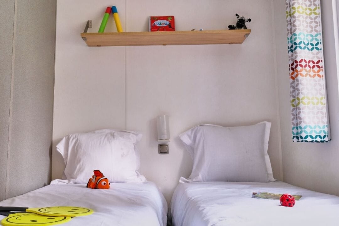 Habitación de hotel con dos camas, ropa de cama blanca, un estante con juguetes y una cortina estampada.