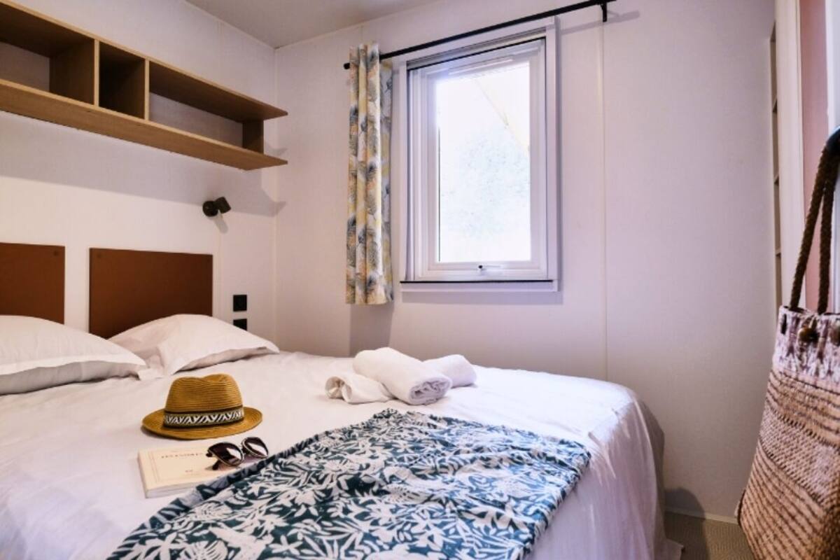 A neatly made bed with a patterned blanket, a hat, sunglasses, and towels on a white wall with a window and a shelf above it.