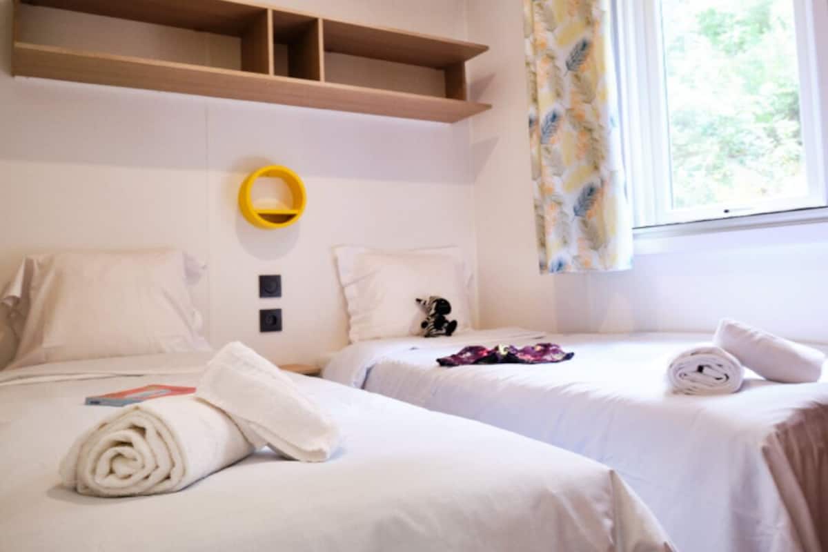 A hotel room with two beds, a wooden shelf above the beds, a yellow wall-mounted object, and a window with curtains.
