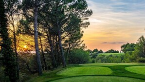 Um campo de golfe com vista para o pôr do sol, rodeado por árvores altas e zonas relvadas.