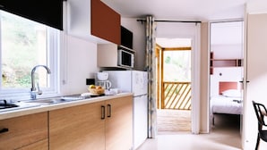 Cozinha moderna com armários de madeira, micro-ondas e pia. Há uma varanda com guarda-corpo de madeira e uma cama visível através de uma porta aberta.