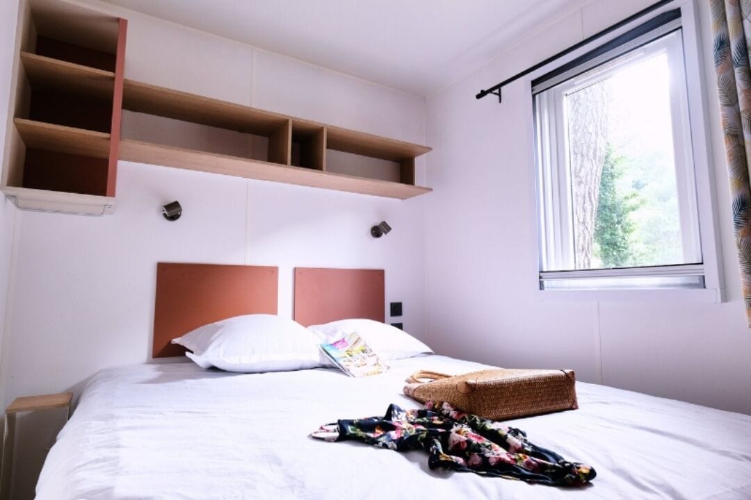 A neatly made bed with a headboard, a window with curtains, and built-in shelves above the bed.